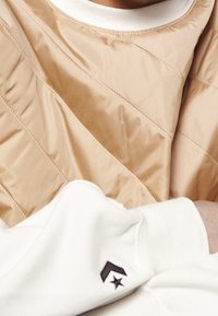 Chaleco acolchado beige con costuras diagonales, superpuesto a una sudadera blanca, que presenta un logo bordado en negro en la manga.