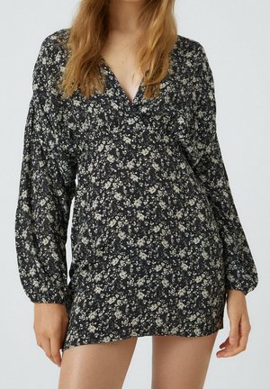 Femme portant une mini-robe noire à manches longues avec un imprimé floral beige et un décolleté en V, sur un fond uni.