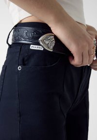 Czarne dżinsy o tkanej fakturze, z klamrą paska w kolorze srebra i złota oraz białą metką "Guess Jeans".