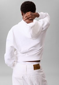 Sudadera blanca corta con capucha y mangas largas, usada sobre unos vaqueros blancos. Se observa la notable cinturilla de Calvin Klein. Textura simple y lisa.