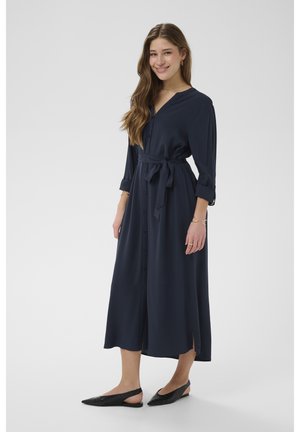 Smaidoša sieviete ar gariem matiem, tērpusies tumši zilā vidēja garuma kleitā ar pogām, uzrullētām piedurknēm un piesietu jostu, melnas slingbeka kurpes un zelta rotaslietas.