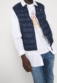 Hombre con chaleco acolchado azul marino sobre camisa blanca de botones y camiseta gráfica, combinado con jeans azules, sobre fondo liso.