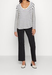Vrouw in een witte blouse met lange mouwen met zwarte horizontale strepen, zwarte rechte broek en beige blokhak schoenen.