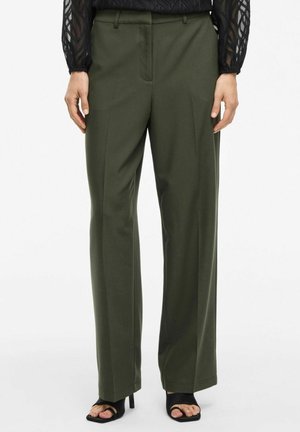 Pantalon vert taille haute droit porté avec des talons ouverts noirs et un top noir transparent à manches longues.