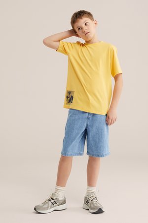 Fant v rumeni majici s potiskom palm, modrih jeansa kratkih hlač, belih nogavic in sivih superg stoji z eno roko za glavo.