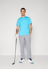 Hellblaues, feuchtigkeitsableitendes Golfshirt mit kurzen Ärmeln, kombiniert mit grauen Golfhosen. Das Modell hält einen Golfschläger und trägt weiße Schuhe mit blauen Akzenten.