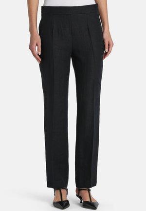 Woman wearing straight-leg black trousers with front pleats and black pointed-toe T-strap heels, standing against a white background.