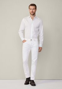 Bílá košile s button-up zapínáním a slim fit střihem, v kombinaci s bílými džínami a tmavě hnědými botami, vystavená na neutrálním pozadí.