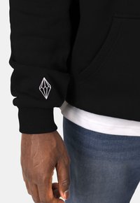 Sweat à capuche noir avec des poignets côtelés, arborant un logo brodé en forme de diamant blanc sur la manche droite, porté par-dessus une chemise blanche.