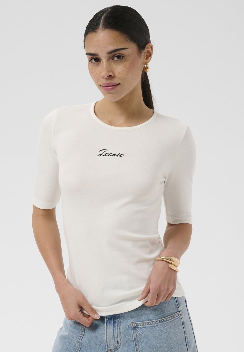 Femme aux cheveux foncés en queue de cheval, portant des boucles d'oreilles créoles dorées, un t-shirt blanc avec le texte « Iconic », un jean bleu et des bracelets dorés, posant sur un fond uni.