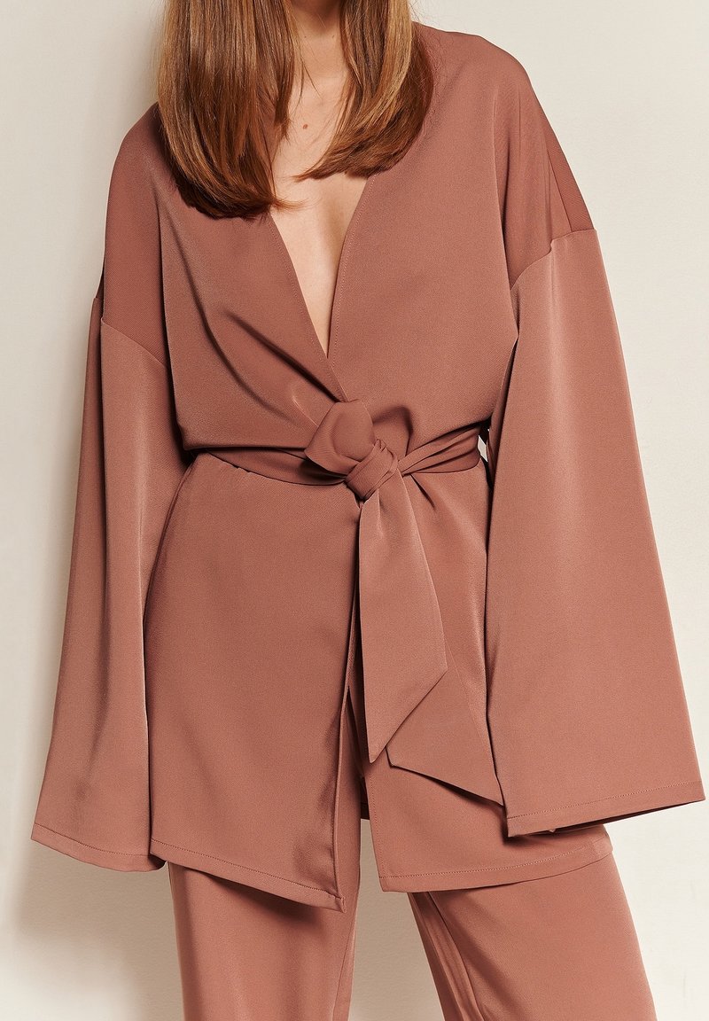 Woman wearing a muted brown satin outfit with wide sleeves and a tied belt at the waist, standing against a plain light wall.