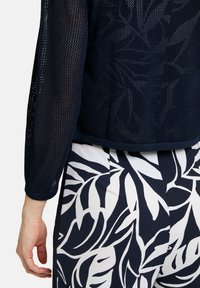 Marineblauer, gestrickter Pullover mit langen Ärmeln und geripptem Saum, kombiniert mit gemustertem weißen und marineblauen Stoff mit tropischen Blättern.