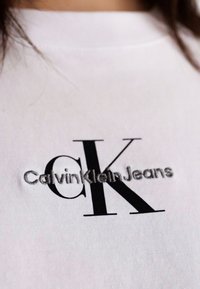 Witte katoenen t-shirt met een zwart en zilver geborduurd logo: "CK" in grote letters en "CalvinKleinJeans" in kleinere tekst eronder.