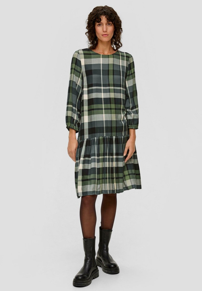Grünes karriertes Kleid mit drei Viertel Ärmeln, gerafftem Taillenbund und gestuftem Rock. Kombiniert mit schwarzen Knöchelstiefeln und transparenten schwarzen Strumpfhosen.