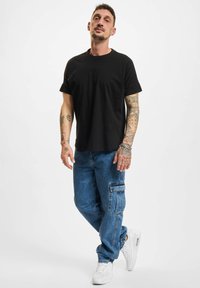 Schwarzes Baumwoll-T-Shirt mit kurzen Ärmeln, kombiniert mit hellblauen Denim-Cargohosen mit einer Seitentasche und weißen Sneakern.