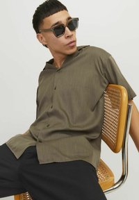 Žalios spalvos, trumpomis rankovėmis, marškiniai su sagomis, laisvu kirpimu, su stovinčiu apykakle ir tamsiomis sagsčiais, dėvimi ant juodų kelnių.