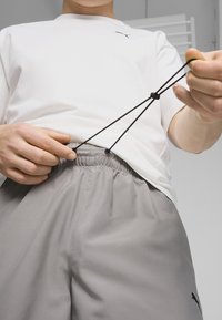 Camisa blanca de manga corta y pantalones cortos de cintura elástica grises con cordón negro. Logo visible en la camisa y en los pantalones. Textura de tela suave.