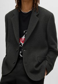 Blazer di un grigio scuro con texture, chiusura a un pulsante, rever a tacche e tasche laterali, indossato sopra una t-shirt nera con grafica.