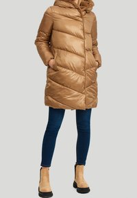 Vrouw in een lichtbruine gewatteerde lange pufferjas met capuchon, blauwe skinny jeans en lichtbruine enkellaarzen met zwarte zolen.