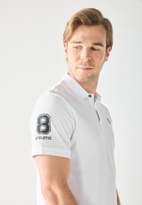 Polo shirt blanc avec un « 8 » noir et une broderie « ATHLETIC » sur la manche gauche. Col classique et patte de boutonnage à deux boutons. Texture lisse.
