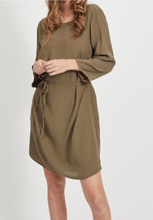 Vrouw draagt een olijfgroene jurk tot op de knie met lange mouwen en een strikceintuur, staand tegen een effen witte achtergrond.