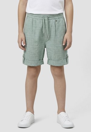 Niño con pantalones cortos verde claro con puños doblados, cintura elástica y cordón, combinados con zapatillas blancas y camiseta blanca.