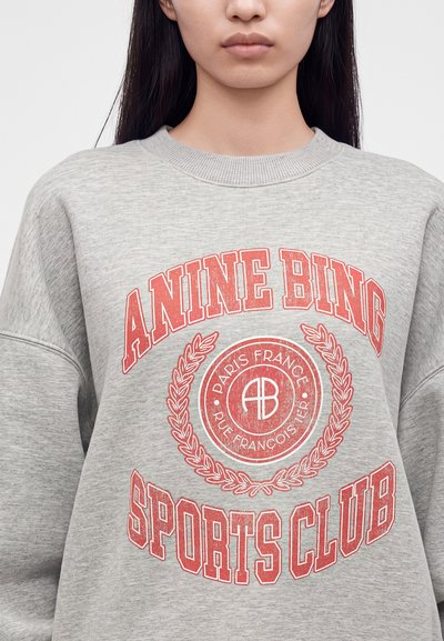 Jeune personne aux longs cheveux foncés portant un sweat gris avec un texte rouge indiquant "Anine Bing Sports Club" et un logo circulaire au centre.