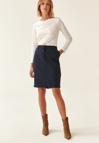 Jupe en coton marine avec taille à cordon, longueur genoux et poches latérales, associée à un haut ajusté à manches longues blanc et des bottines brunes.