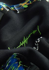 Közeli kép fekete anyagról neon zöld absztrakt varrással és sokszínű, kék, zöld és sárga festékfröccsenésekkel.