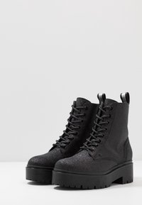 Bottines noires pailletées à lacets avec semelles compensées épaisses et languettes à l’arrière, posées côte à côte sur une surface blanche.