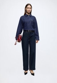 Camisa de ganga de manga comprida com colarinho, jeans azul-escuro de cintura alta e perna larga, e uma mala vermelha de couro. Sapatos pretos de bico fino completam o visual.