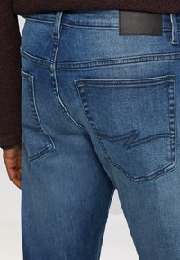 Un jean en denim bleu foncé présentant un gros plan de l'arrière avec deux poches plaquées et un design cousu. Il dispose d'une étiquette de marque en cuir au niveau de la taille.