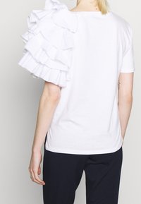 Valkoinen puuvillainen t-paita, jossa on röyhelöillä koristellut olkapäät. Tasainen pinta, pyöreä pääntie ja lyhyet hihat. Yksinkertainen, koristeeton takaosa.