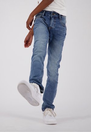Jean en denim bleu délavé avec une coupe slim, un design à cinq poches et une fermeture par bouton. Porté avec des baskets blanches.