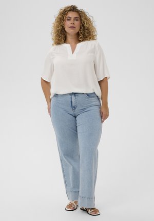 Woman with curly hair wearing a white blouse, light blue jeans, and white sandals standing against a plain background.