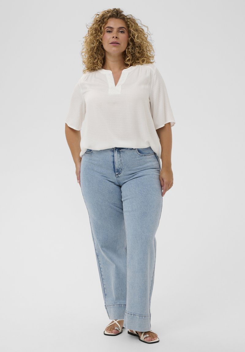 Femme aux cheveux bouclés portant une blouse blanche, un jean bleu clair et des sandales blanches, debout devant un fond uni.