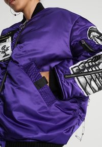 Personne portant une veste bomber violette brillante avec divers patchs noirs et blancs et des poches zippées sur les manches et la poitrine.