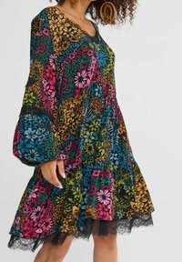 Femme portant une robe patchwork florale colorée avec de longues manches ballons et un ourlet bordé de dentelle, accessoirisée de grandes boucles d'oreilles circulaires.