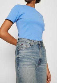 Person iført en tætsiddende lyseblå kortærmet skjorte, der er stukket ned i højtaljede falmede blå jeans med et lille rød, hvidt og blåt mærke på lommen.
