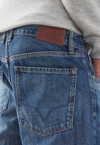 Blå denimjeans med en brun läderetikett som har "Pepe Jeans". Designen inkluderar två bakfickor och kontrasterande sömmar.