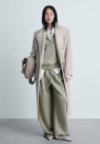 Beige dubbelzijdige jas over een lichtgroene sweatshirt en bijpassende brede broek. Accessoire: zachte beige handtas. Platte, puntige schoenen.