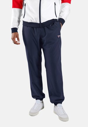 Man draagt marineblauwe Reebok trainingsbroek, witte sneakers en een witte jas met rode en marineblauwe accenten.