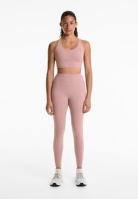  Lichtroze sport-bh en bijpassende high-waisted leggings. De stof lijkt glad met een nauwsluitend ontwerp en minimale naden. Witte sportschoenen.