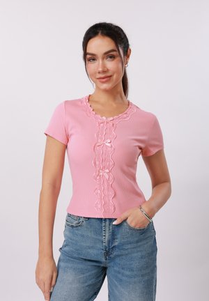 Ung kvinde iført en tætsiddende lyserød kortærmet top med blonder og blå jeans, poserer med den ene hånd i lommen mod en ensfarvet baggrund.