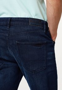 Mörkblå denimjeans har en klassisk femficksdesign med en läderlogotyp på midjebandet och subtila utslitage detaljer.