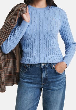 Personne portant un pull en maille torsadée bleu clair et un jean bleu, tenant un manteau marron à carreaux sur une épaule.
