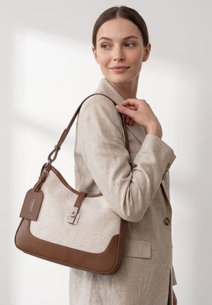 Vrouw in beige blazer houdt een beige en bruine leren schoudertas met zilveren gesp vast, kijkt over haar schouder tegen een effen achtergrond.