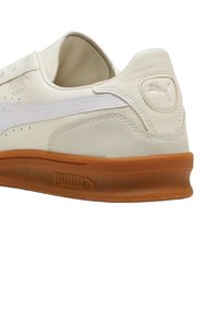 Sneaker color crema con tomaia in pelle liscia, striscia bianca laterale e suola in gomma marrone a contrasto; presente logo in rilievo sul tallone.