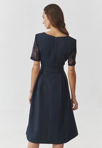 Marineblaues Kleid mit taillierter Taille, kurzen Spitzenärmeln, Reißverschluss auf der Rückseite und einem ausgestellten Rock. Glatte Textur mit klassischer Silhouette.