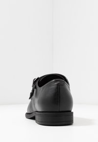 Chaussure en cuir noir vue de l'arrière, dotée d'une boucle décorative et d'une surface lisse, sur fond blanc.